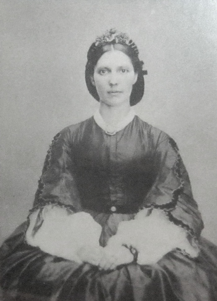 Black and white photograph of young woman in dark dress with arms folded in her lap, hair tied in a bun beneath.