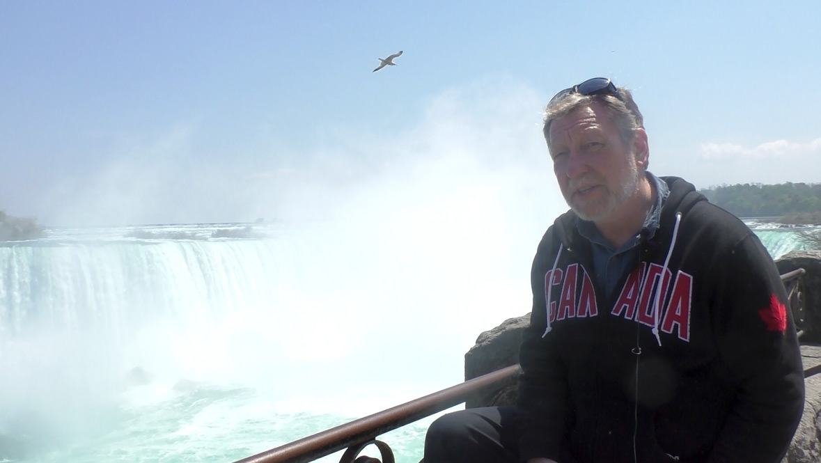 Man with beard and dark blue sweatshirt with 'Canada' inscribed in red faces camera at railing of Niagara Falls.