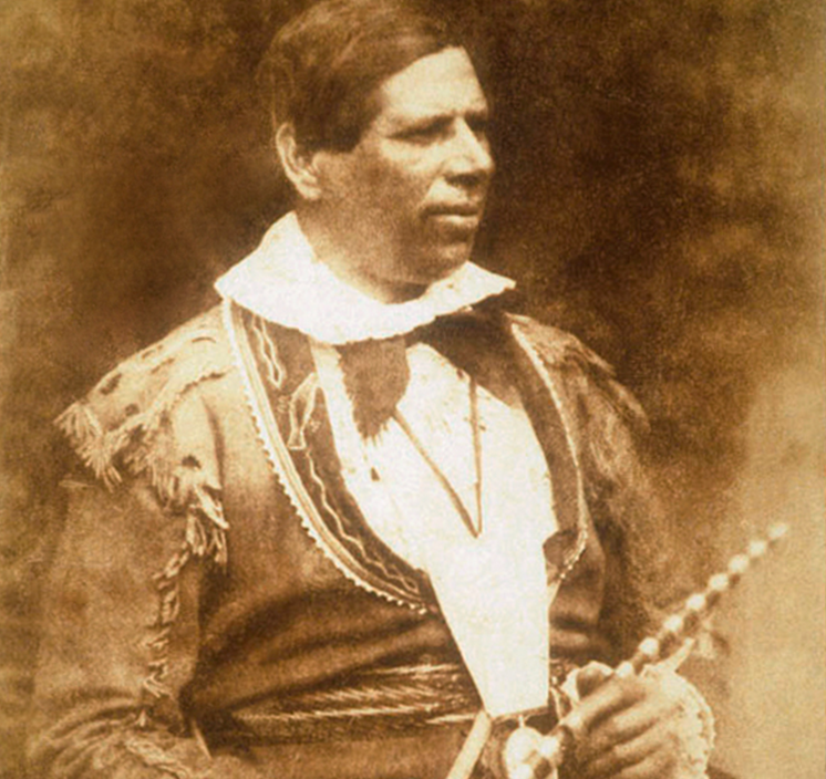 Photographie en noir et blanc d’un homme d’âge mûr, aux cheveux foncés et aperçu de profil, à partir de la taille, habillé de vêtements traditionnels d’un peuple autochtone du Canada et tenant un calumet de la paix dans ses mains.