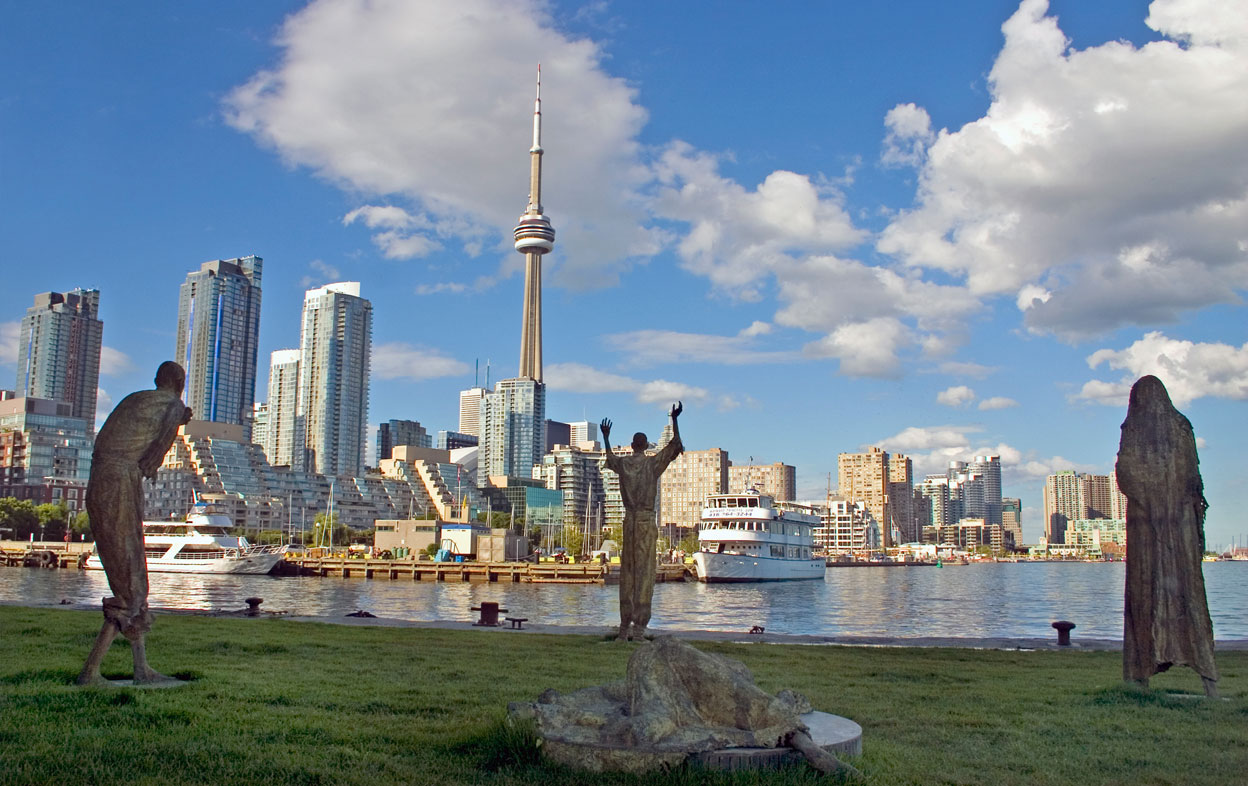 Sculpture of figure lying down in foreground, surrounded by three sculptural figures standing in middle ground, including male figure with arms upraised facing CN Tower and city skyline across an expanse of water. Two white yachts in background.