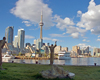 Sculpture of figure lying down in foreground, surrounded by three sculptural figures standing in middle ground, including male figure with arms upraised facing CN Tower and city skyline across an expanse of water. Two white yachts in background.