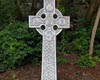 Grey Grey Celtic Cross with ornate patterning viewed from front, with brown earth and green foliage in background.