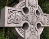 Slightly diagonal view of top of  grey Celtic Cross with icons clockwise from of pick and shovel, mosquito, wheelbarrow, and explosion.