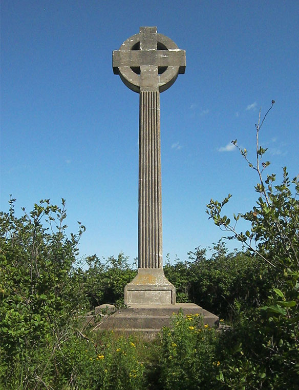 Tall grey Celtic Cross with blue sky in background, lush vegetation at its base.