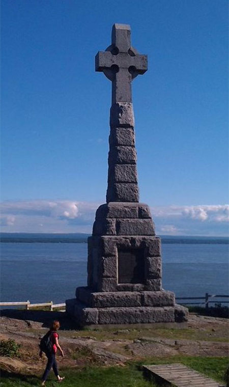 Une croix celtique et une personne s’y approchant, avec le fleuve et un ciel bleu aperçus à l’arrière-plan.
