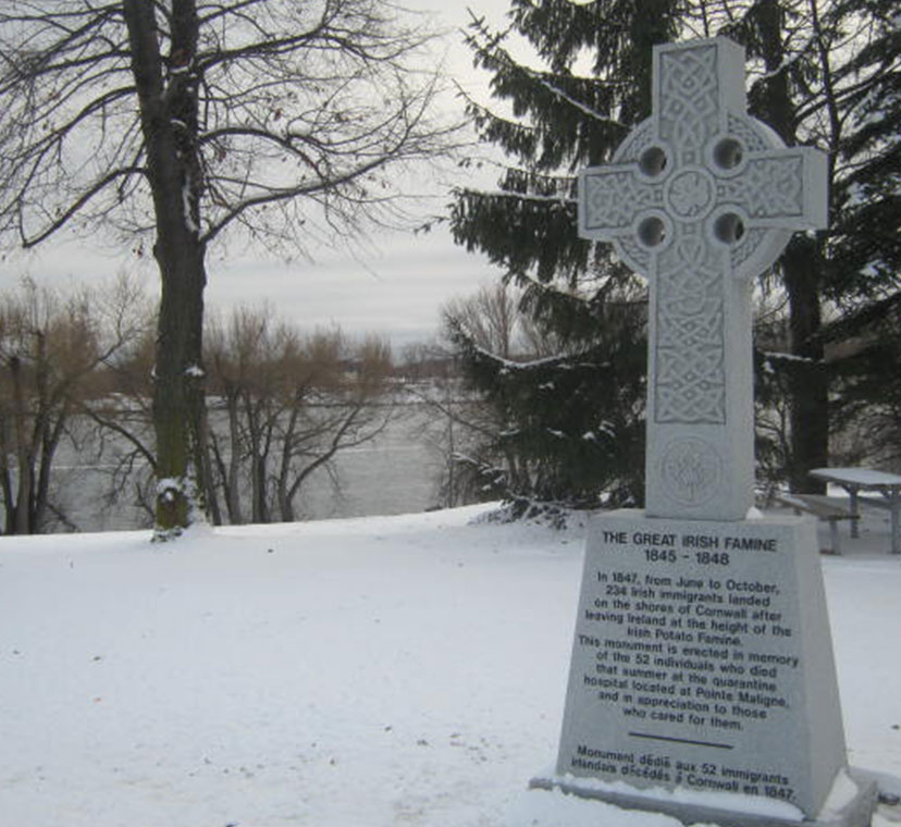 Une croix celtique de couleur grise, aperçue de biais sur un terrain enneigé et près d’une rivière. Il y est inscrit, sur sa base : La Grande Famine irlandaise, 1845-1848, ainsi que d’autres informations en plus petits caractères.