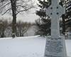 Grey Celtic Cross viewed diagonally in snowy ground, beside river. Inscribed on its base: The Great Irish Famine. 1845-1848, with additional smaller inscriptions underneath.