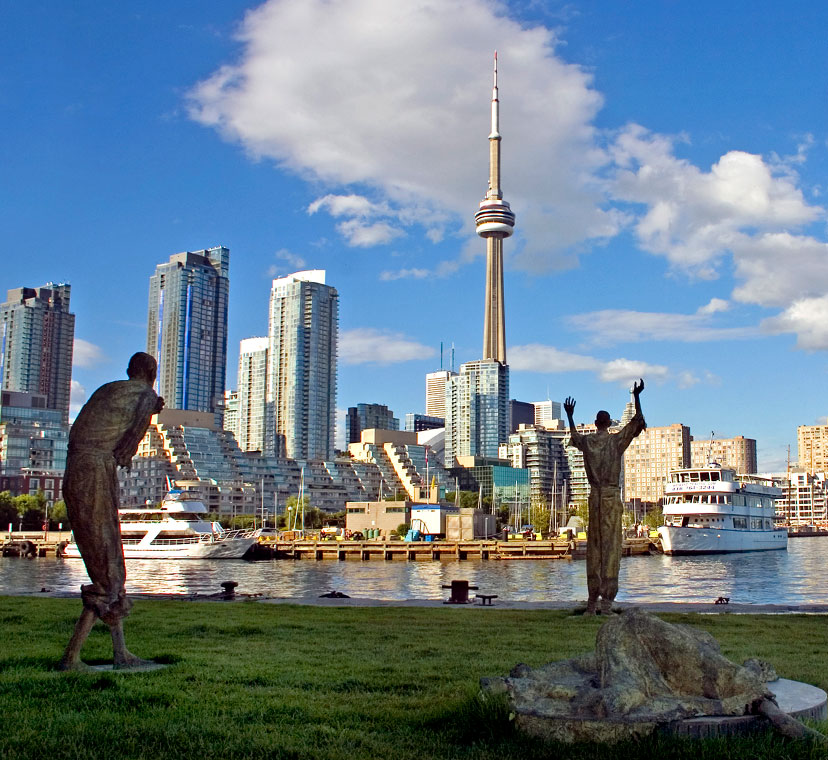 Wide angle image with one sculptural figure lying down in foreground, three standing sculptural figures in middle ground, and city skyline and CN Tower in background across an expanse of water, under a blue sky with white clouds.