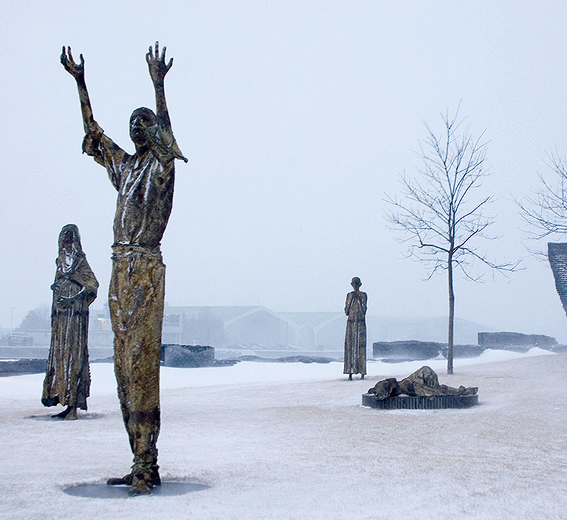 Wide angle image with five sculptural figures, sculpture of man standing with arms upraised in foreground with pregnant woman clutching her stomach to his left, dying woman lying down behind him to his right. Sculpture of man hunched over further right, and sculpture of a boy clutching his hands to his chest in middle ground left. Winter scene with snow on ground, several trees with no leaves, and sculptural wall in background.