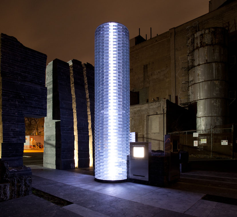 Beacon of light single column of glass lit up at night, surrounded by sculptural stone wall in background. Stone floor stone slab tiles in foreground.