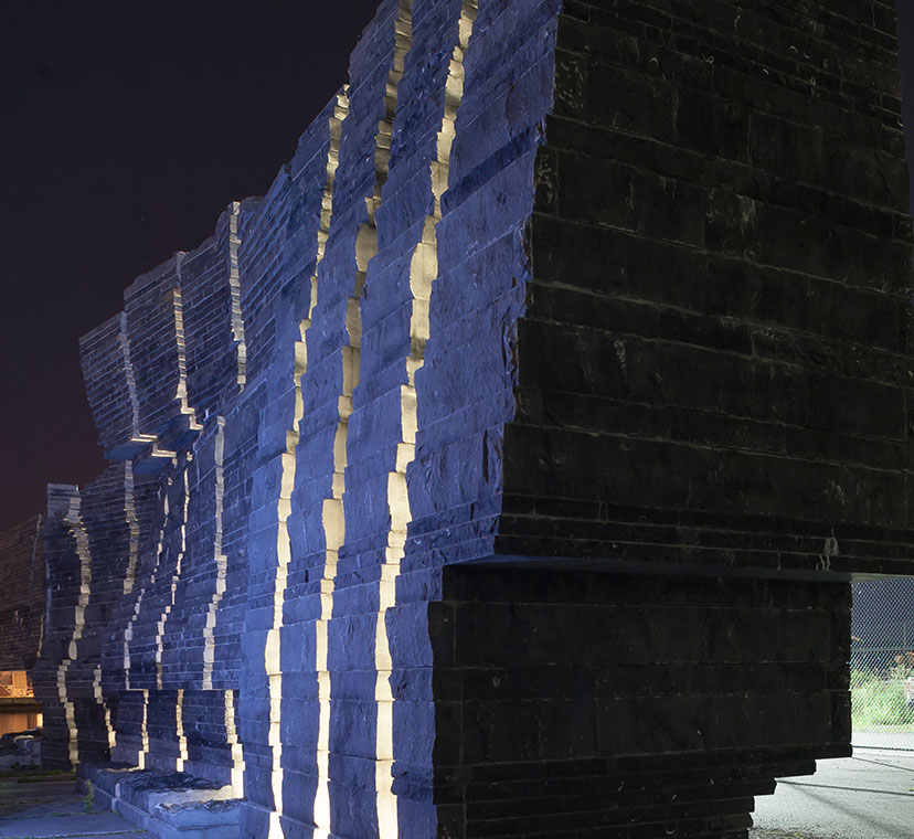 Vue diagonale du mur de pierres sculptées, illuminé, aux blocs de pierre qui sont posés à sa base.