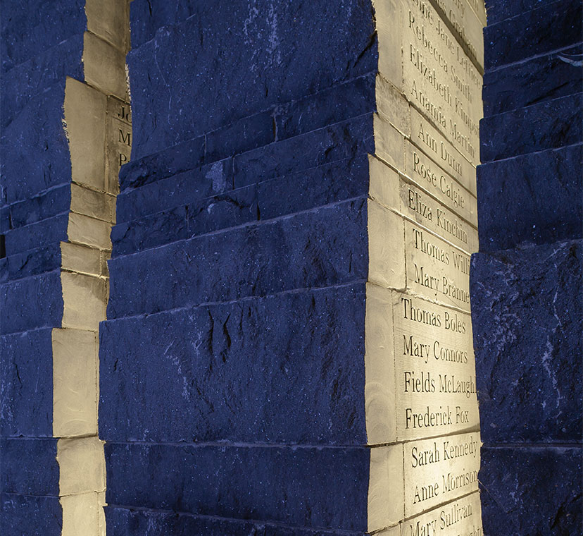 Irregular shaped stone column with names engraved on right diagonal side, backlit at night.