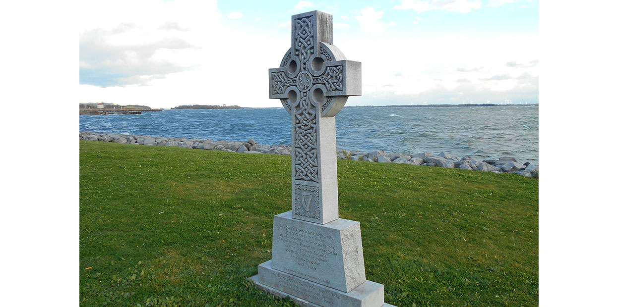 Diagonal view of grey Celtic Cross with ornate patterning and inscription on plinth, on green grass with stones on shore, choppy water, green land masses and blue cloudy sky visible in background.
