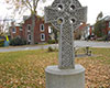 Small grey Celtic Cross with ornate patterning on circular plinth, with leaves on ground, road, and dark red coloured houses in background.
