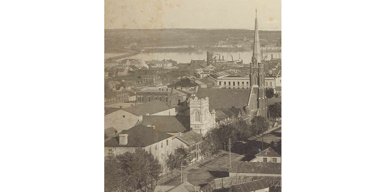 Black and white photo of street from above, first three storey building on left in three quarters profile, adjacent to small church with square tower, four houses, and much larger church with soaring steeple, city skyline and lake in background.