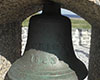 Grey bell mounted in monument with Carricks 1823 inscribed on it. Grey brick floor and wall, grass, and the sea visible in background.