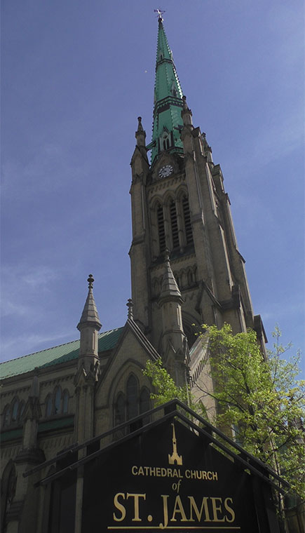 Cathedral steeple and front facade of church, with leafy tree and sign for Cathedral Church of St James in foreground. Blue sky in background.