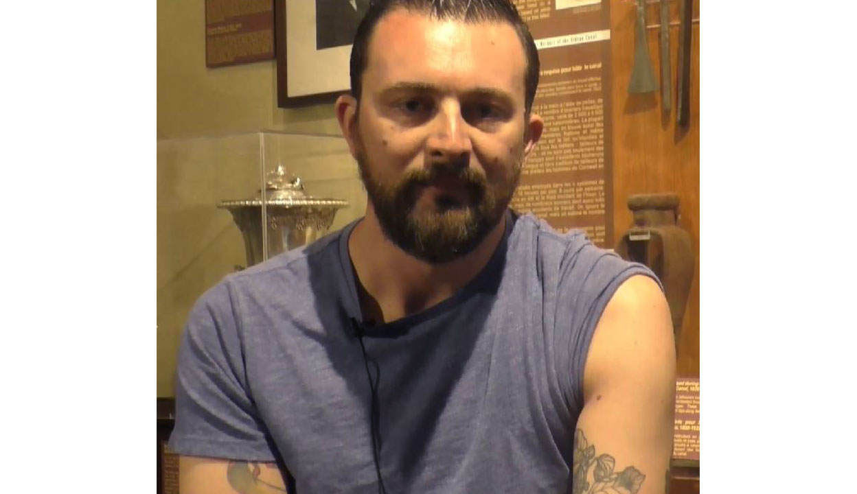 Colour photograph of a young man with short brown hair, moustache, and beard looking to camera. He is wearing a light blue tee-shirt and tattoos are visible on both of his arms below the sleeves. Behind him can be seen a silver chalice in a glass display case and a brown interpretive board.