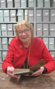 Smiling woman in glassed holding open brown diary, seated at brown table. Numerous grey archive folders shelved in background.